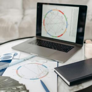 Astrology charts displayed on a laptop next to tarot cards and notebooks on a table.